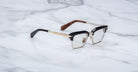 The Jacques Marie Mage Alin eyeglasses feature black and gold frames, clear lenses, and brown patterned temple arms, displayed on a white marble surface.