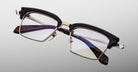 The Jacques Marie Mage Alin features rectangular eyeglasses with a black upper frame, gold accents, clear lenses, and brown patterned temples in a modern, stylish design displayed on a light gray background.
