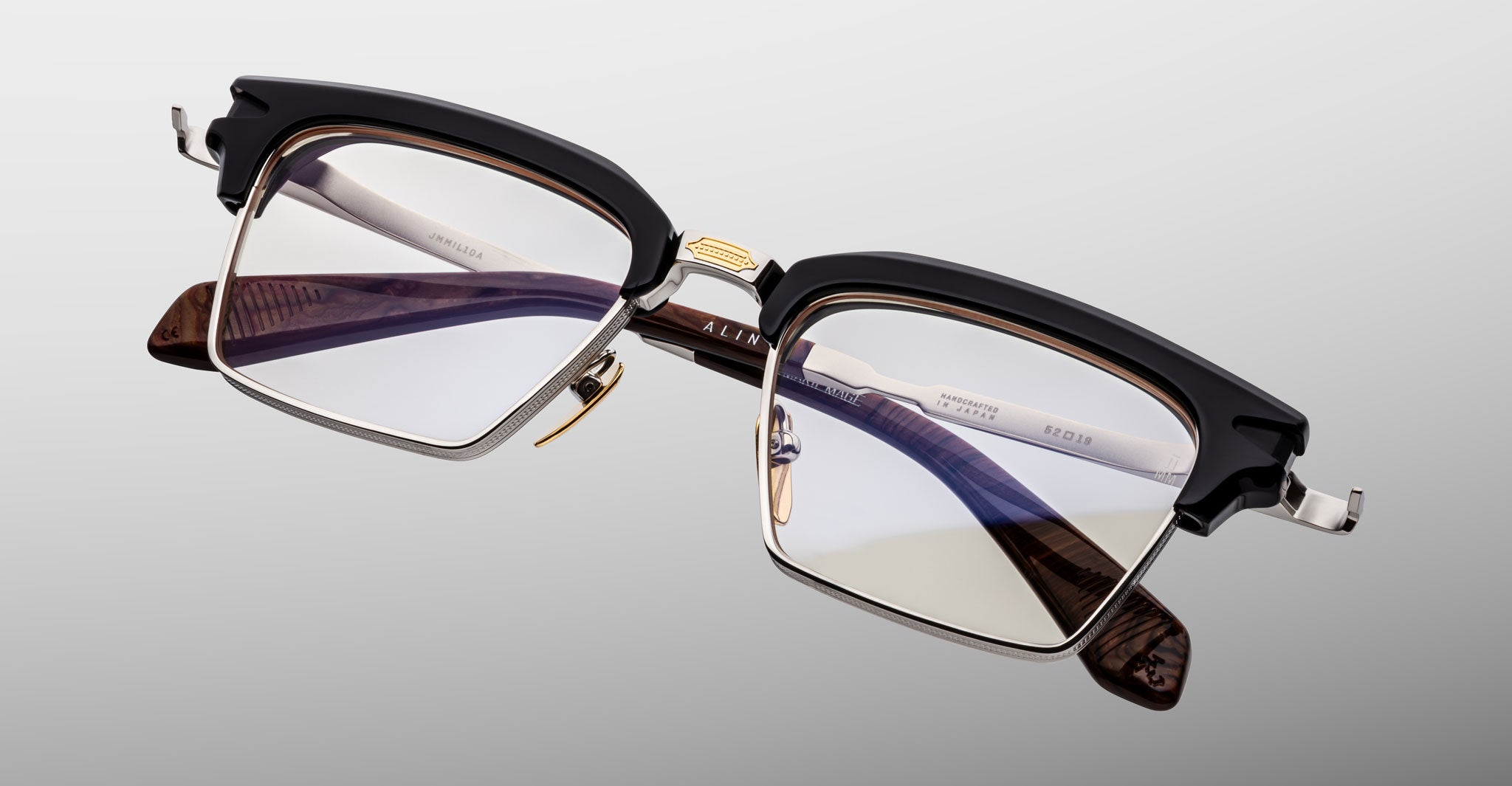 The Jacques Marie Mage Alin features rectangular eyeglasses with a black upper frame, gold accents, clear lenses, and brown patterned temples in a modern, stylish design displayed on a light gray background.