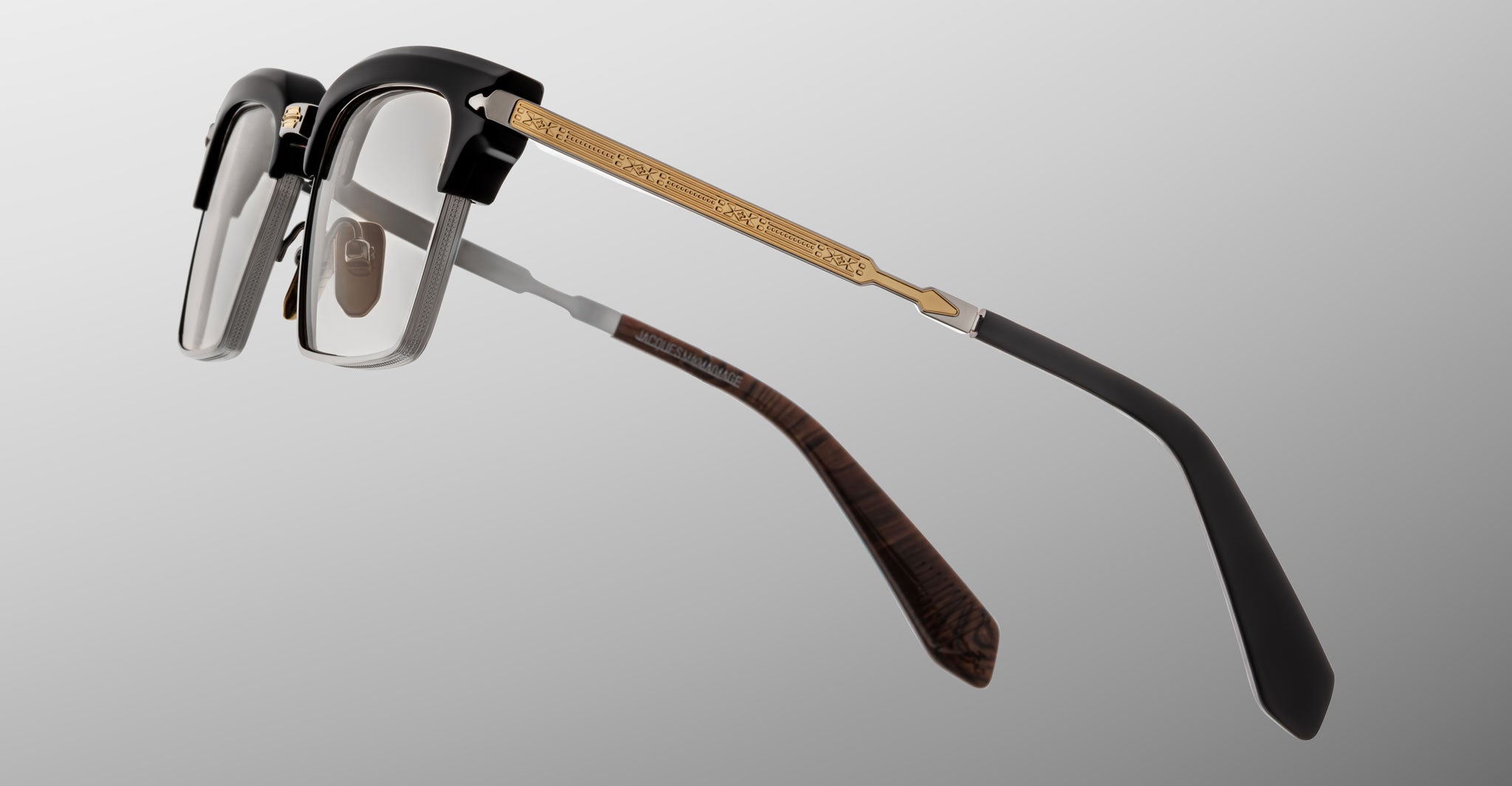 A close-up of Jacques Marie Mage Alin eyeglasses: black frames, gold metal arms with engraved pattern, dark temple tips, and clear lenses set against a light grey gradient background.