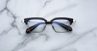 The Jacques Marie Mage Alin, featuring black rims, rectangular lenses, and silver hinges, rests on a white marble surface with gray veining.