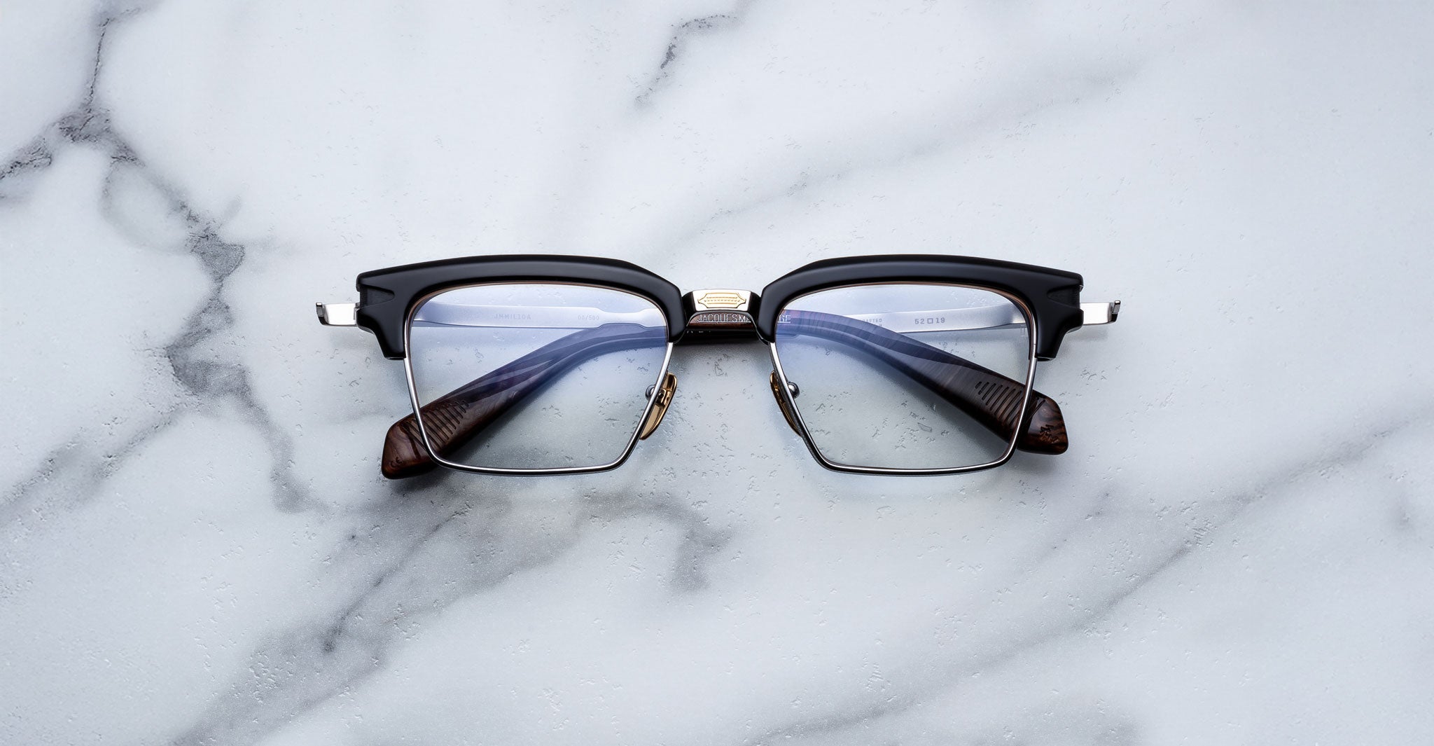 The Jacques Marie Mage Alin, featuring black rims, rectangular lenses, and silver hinges, rests on a white marble surface with gray veining.