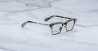 The Jacques Marie Mage Alin features rectangular camouflage-patterned acetate frames, a silver metal bridge and temples, and clear lenses, shown resting on a white marble surface.