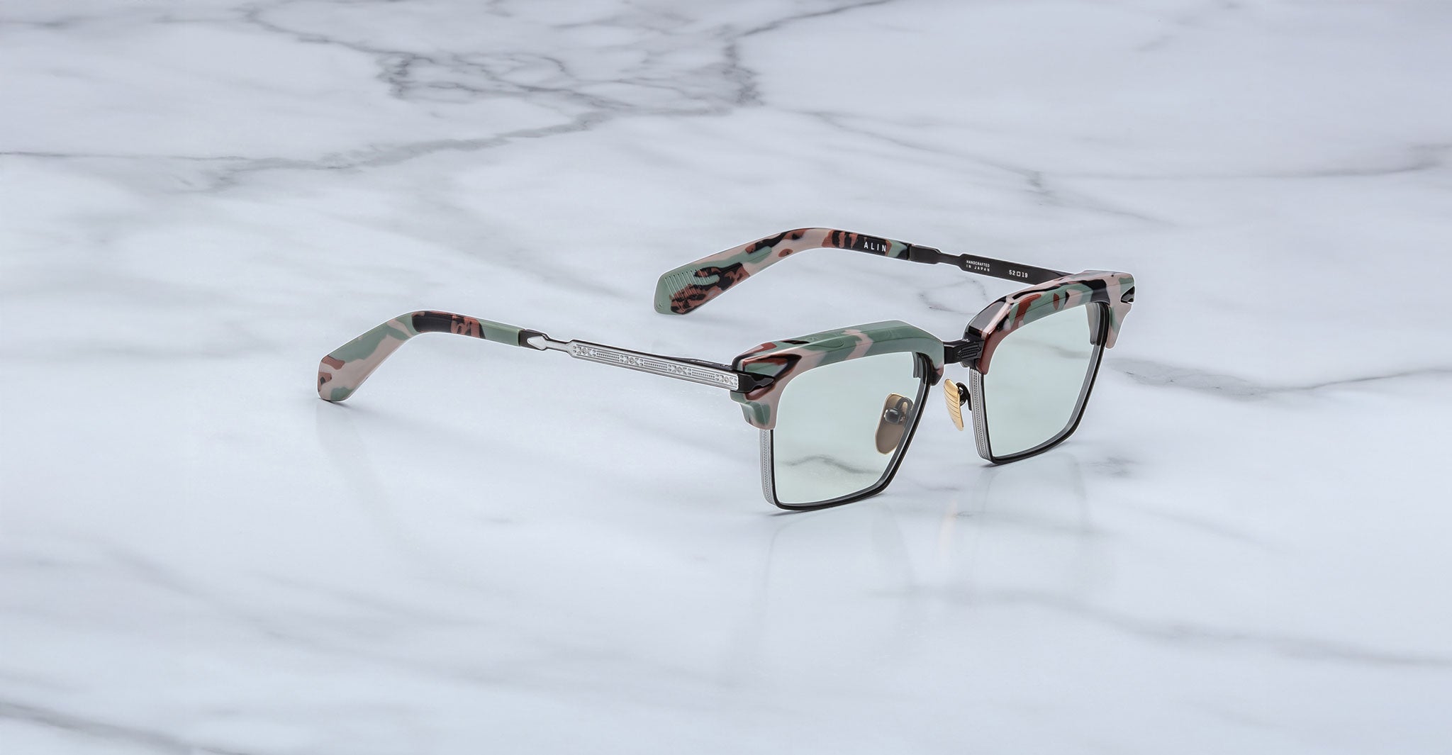 The Jacques Marie Mage Alin features rectangular camouflage-patterned acetate frames, a silver metal bridge and temples, and clear lenses, shown resting on a white marble surface.