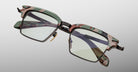 The Jacques Marie Mage Alin eyeglasses feature a green and pink camouflage frame, clear lenses, black lower rims, and metal bridge details, displayed against a light gray background.