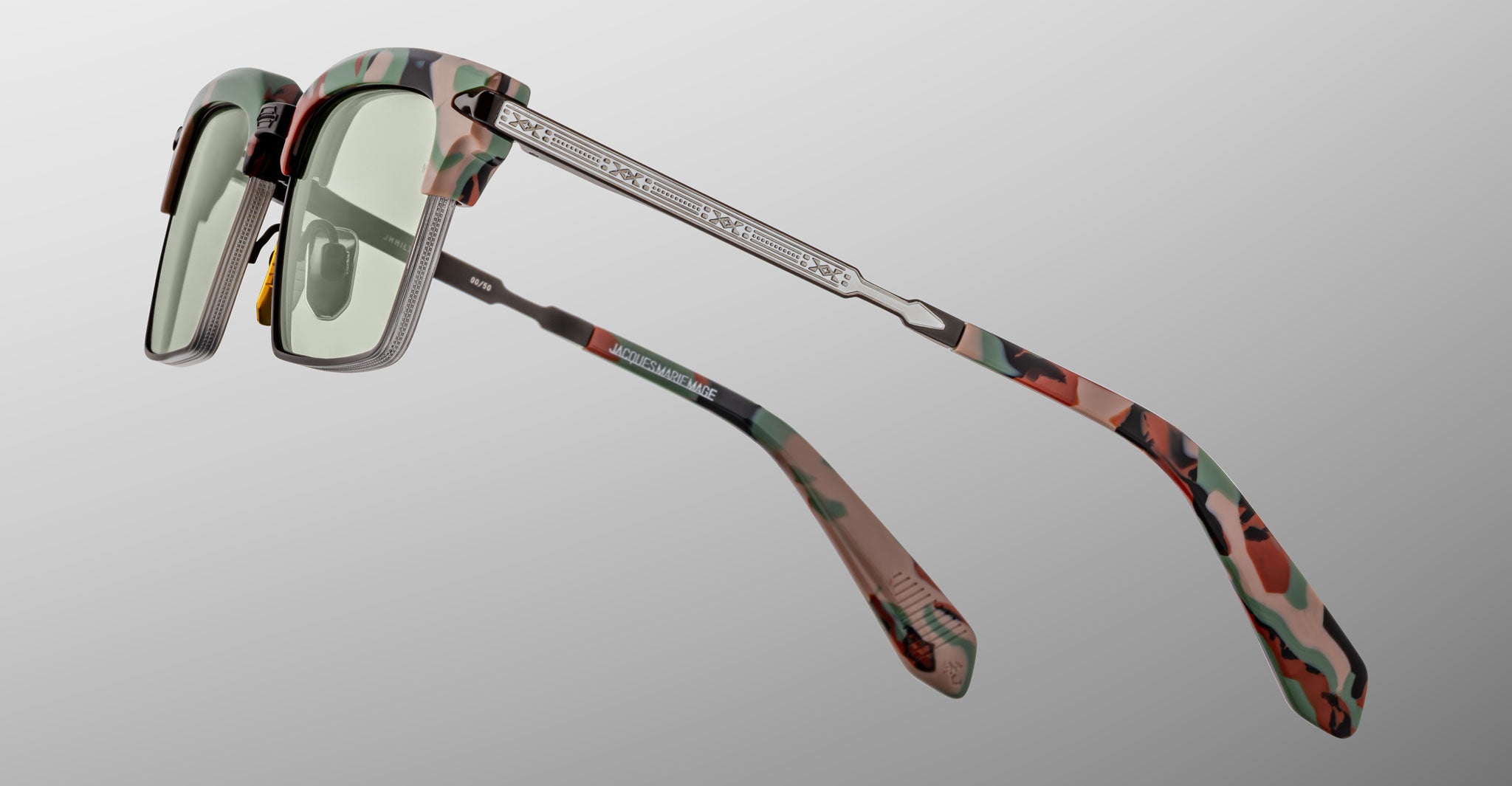 The Jacques Marie Mage Alin sunglasses feature green-tinted lenses, camouflage-patterned frames and arms, and metallic temples with engraved details, showcased against a plain gray background.