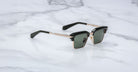 A pair of Jacques Marie Mage Alin black and gold square sunglasses with dark lenses rests on a white marble surface.