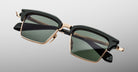 The Jacques Marie Mage Alin features square-shaped sunglasses with dark green lenses, a black upper frame, and gold metal accents, displayed against a light gray gradient background.