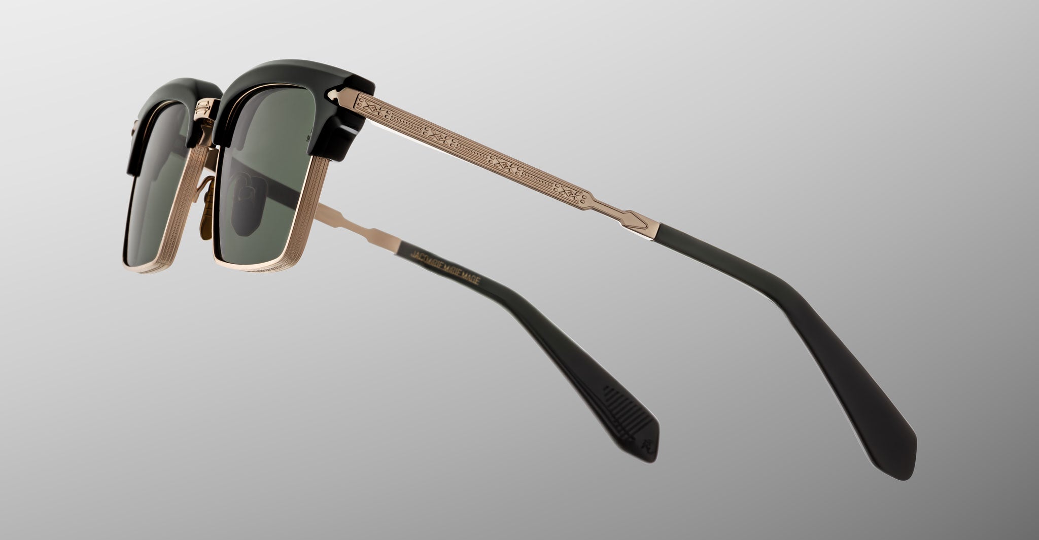 A close-up photo of Jacques Marie Mage Alin sunglasses featuring gold metal frames, dark green lenses, and black temple tips, displayed on a light grey background.