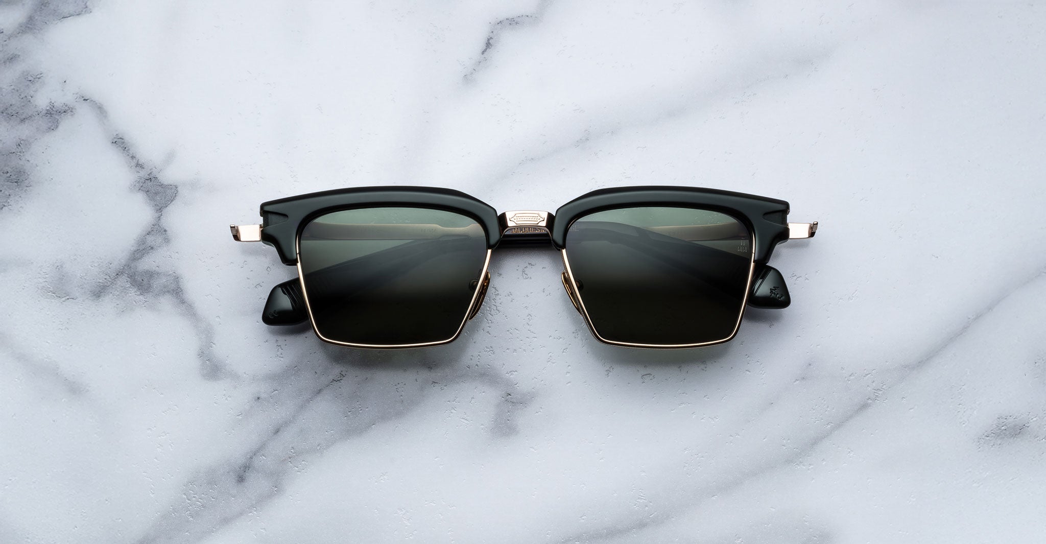 The Jacques Marie Mage Alin sunglasses, featuring black square frames, dark lenses, and gold accents, are displayed on a white marble surface.