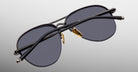 The Jacques Marie Mage Amelia is a pair of black aviator sunglasses featuring dark lenses, gold nose pads, and thin black arms, displayed from the back on a plain gray background.