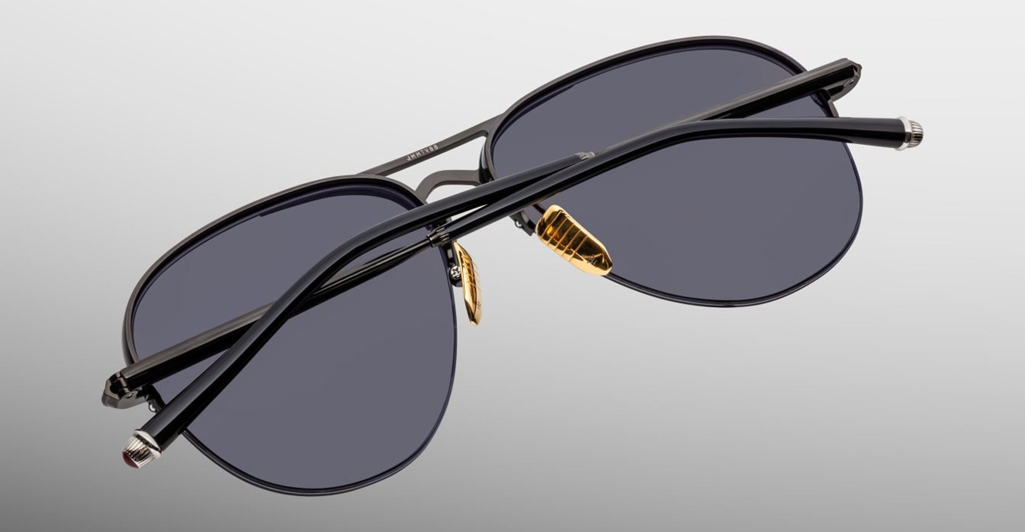 The Jacques Marie Mage Amelia is a pair of black aviator sunglasses featuring dark lenses, gold nose pads, and thin black arms, displayed from the back on a plain gray background.