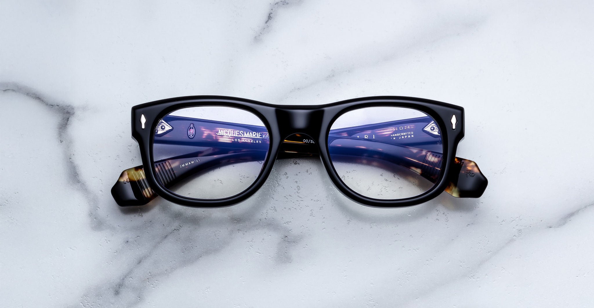 Jacques Marie Mage Amilcar eyeglasses on white marble, cityscape reflected on rims, top-down view.