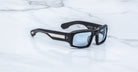 Jacques Marie Mage Apollo in Shadow shown in angled view, featuring a bold rectangular black acetate frame, thick sculpted temples with exposed engraved metal core, triple dot rivets, signature arrowhead tips, and blue-tinted lenses.