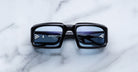 Front view of Jacques Marie Mage Apollo in Shadow featuring a bold rectangular black acetate frame, thick sculpted rims, blue-tinted lenses, and metal hinge details visible through the lens.