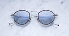 A pair of round, silver-framed sunglasses with blue-tinted lenses rests on a white marble surface. The ends of the temples have small red tips.