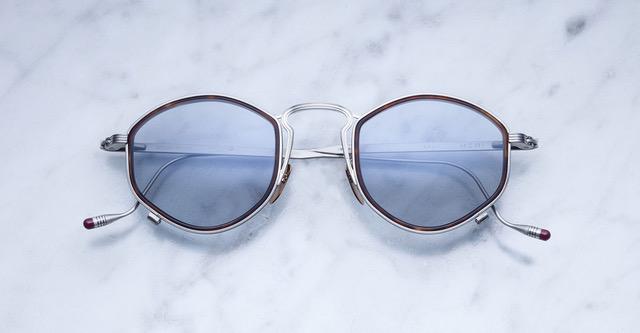A pair of round, silver-framed sunglasses with blue-tinted lenses rests on a white marble surface. The ends of the temples have small red tips.