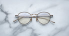 A pair of Jacques Marie Mage Archibald round, Altan light gold-framed optical frame with thin metal arms rests on a white marble surface with grey veining.