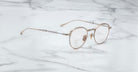 A pair of Jacques Marie Mage Archibald round, Altan light gold-framed optical frame with thin metal arms rests on a white marble surface with grey veining.