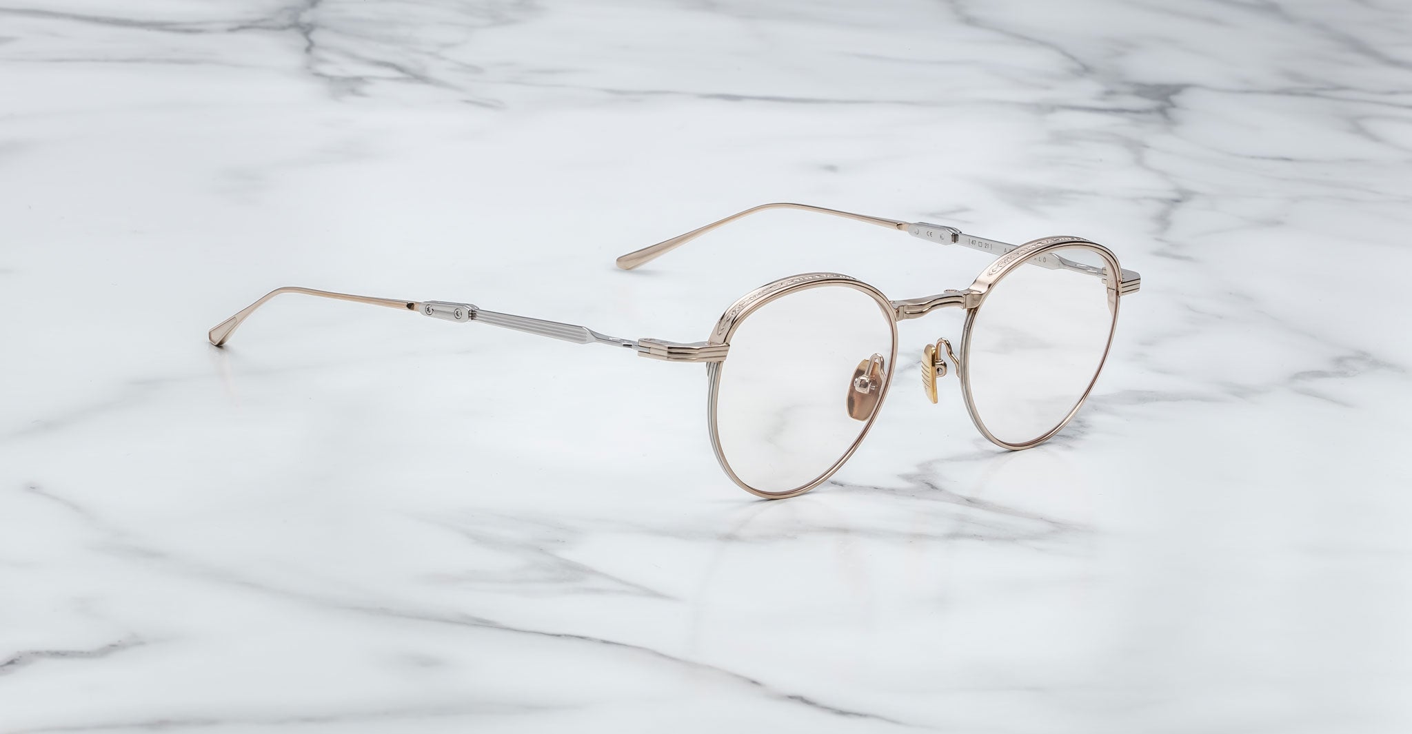 A pair of Jacques Marie Mage Archibald round, Altan light gold-framed optical frame with thin metal arms rests on a white marble surface with grey veining.