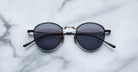 A pair of round, moon gold-framed sunglasses with charcoal lenses is placed on a white marble surface. The arms of the sunglasses are metallic with visible branding on the inner side of the arms.
