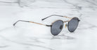 A pair of Jacques Marie Mage Archibald round, moon gold-framed sunglasses with thin metal arms rests on a white marble surface with grey veining.