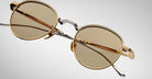 The Jacques Marie Mage Archibald is a pair of round sunglasses featuring gold-colored frames and light brown lenses, displayed at an angle on a plain, gradient gray background.