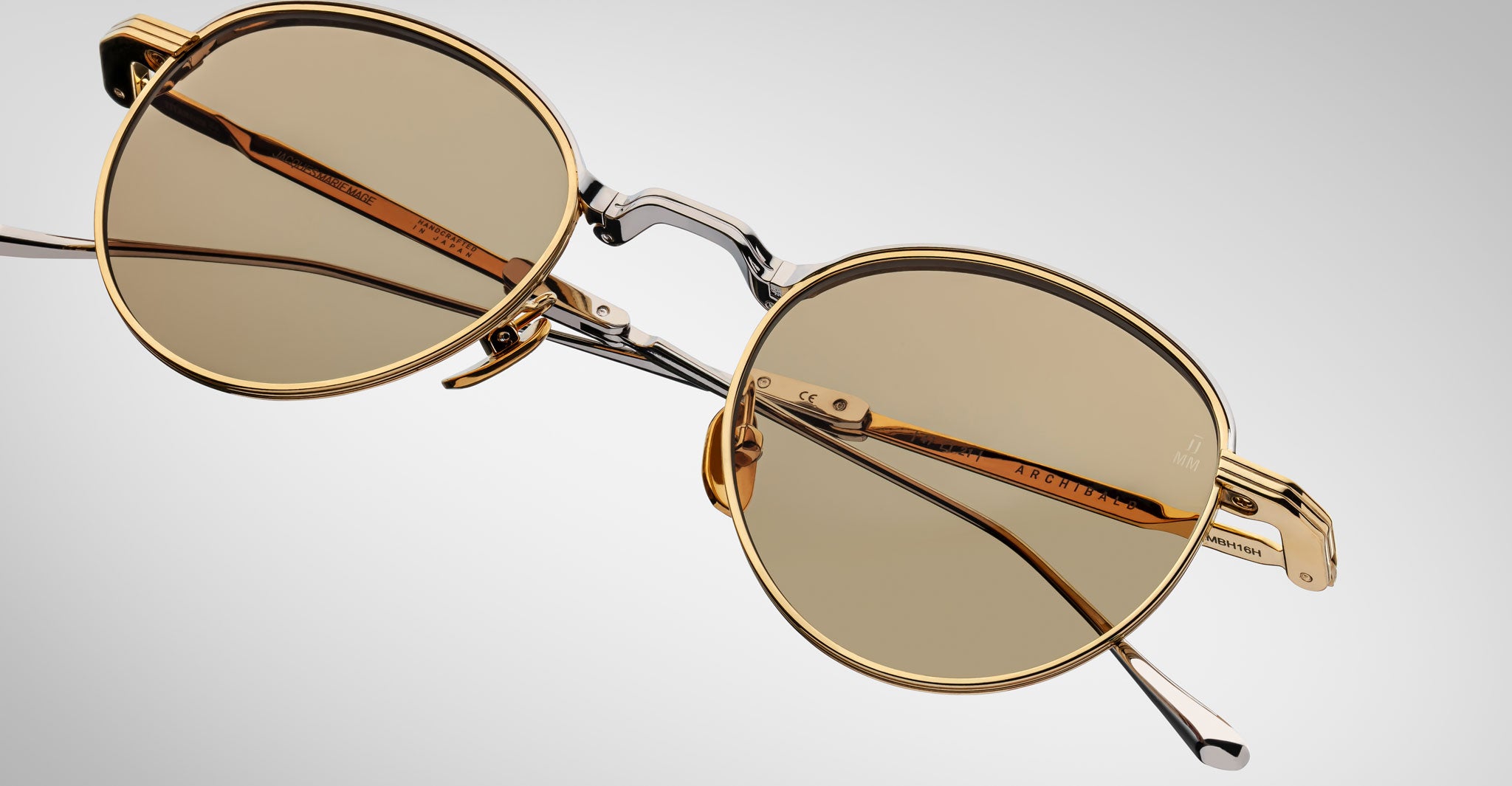 The Jacques Marie Mage Archibald is a pair of round sunglasses featuring gold-colored frames and light brown lenses, displayed at an angle on a plain, gradient gray background.