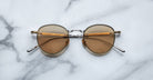 A pair of round, gold-framed sunglasses with brown lenses is placed on a white marble surface. The arms of the sunglasses are metallic with visible branding on the inner side of the arms.