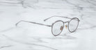 A pair of Jacques Marie Mage Archibald round, silver-framed optical frame with thin metal arms rests on a white marble surface with grey veining.