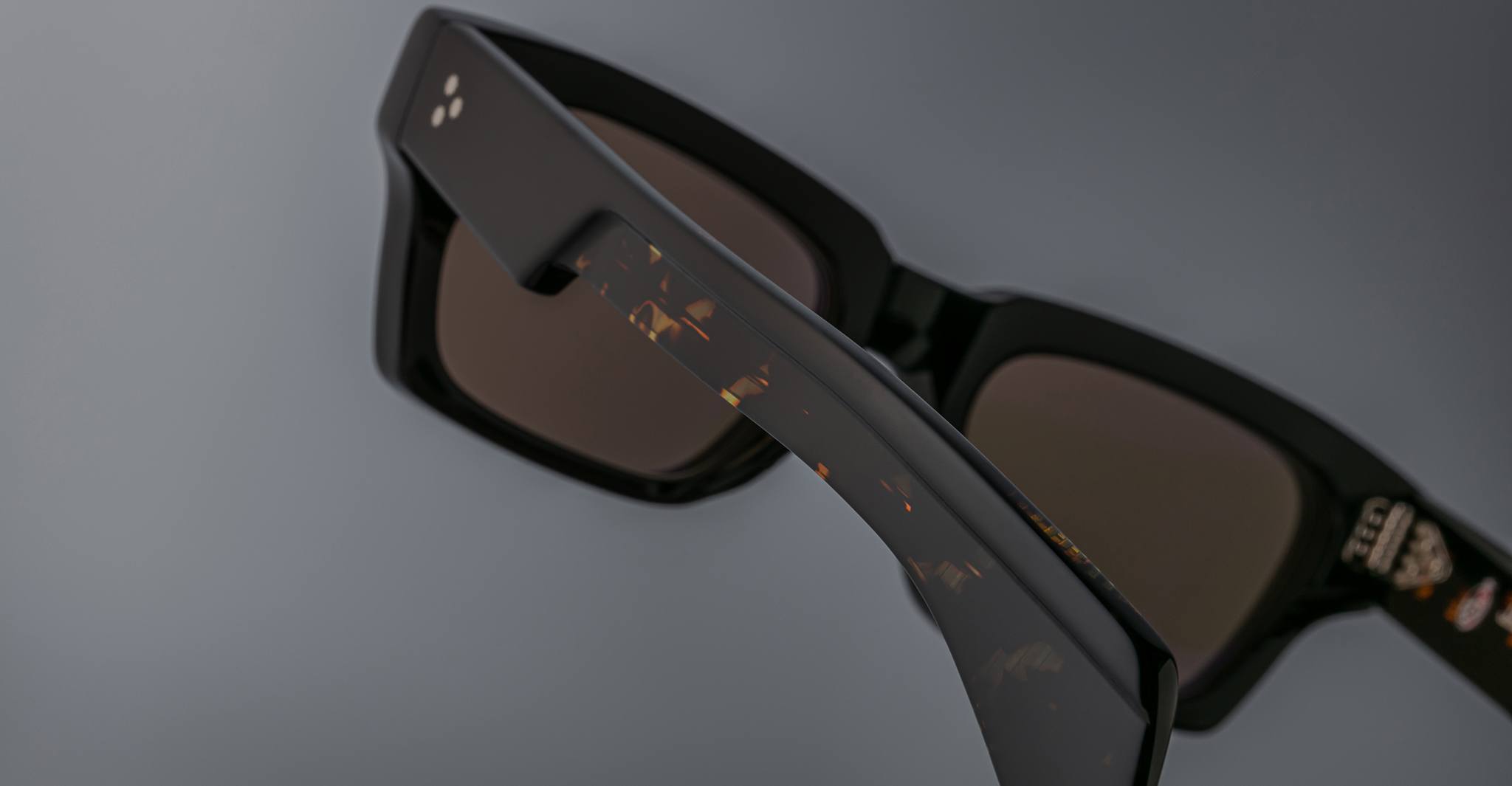Close-up of Jacques Marie Mage Ashcroft black rectangular sunglasses with dark lenses and a partially visible marbled temple pattern, set against a grey background.