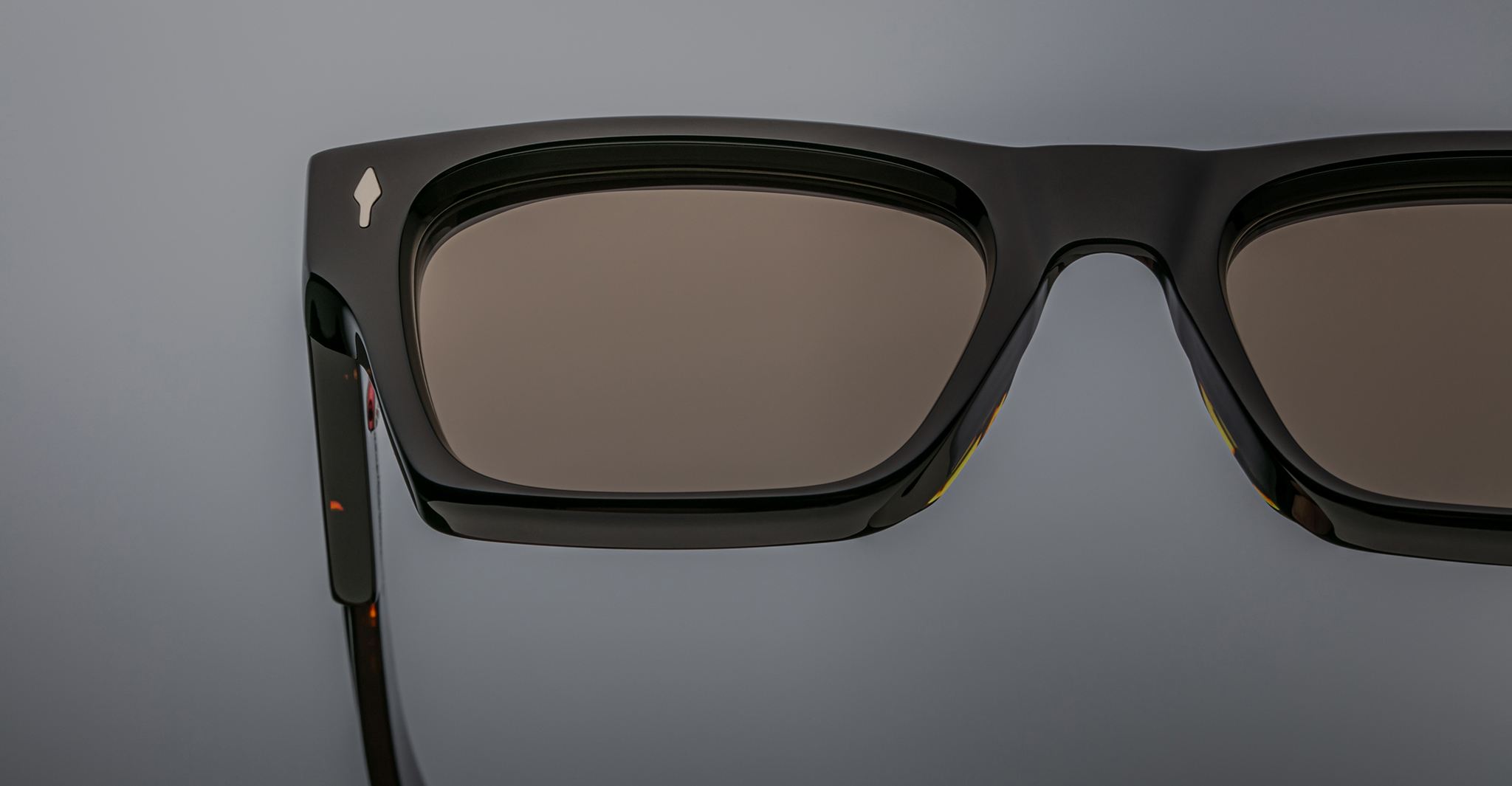 Close-up of Jacques Marie Mage Ashcroft—a pair of black rectangular sunglasses with dark lenses—showing part of the frame and left temple against a neutral gray background.