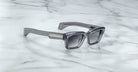 Jacques Marie Mage Ashcroft sunglasses, showcasing gray rectangular thick frames and dark lenses, rest on white marble with gray veining. Metallic accents adorn the arms near the hinges, adding a refined touch.