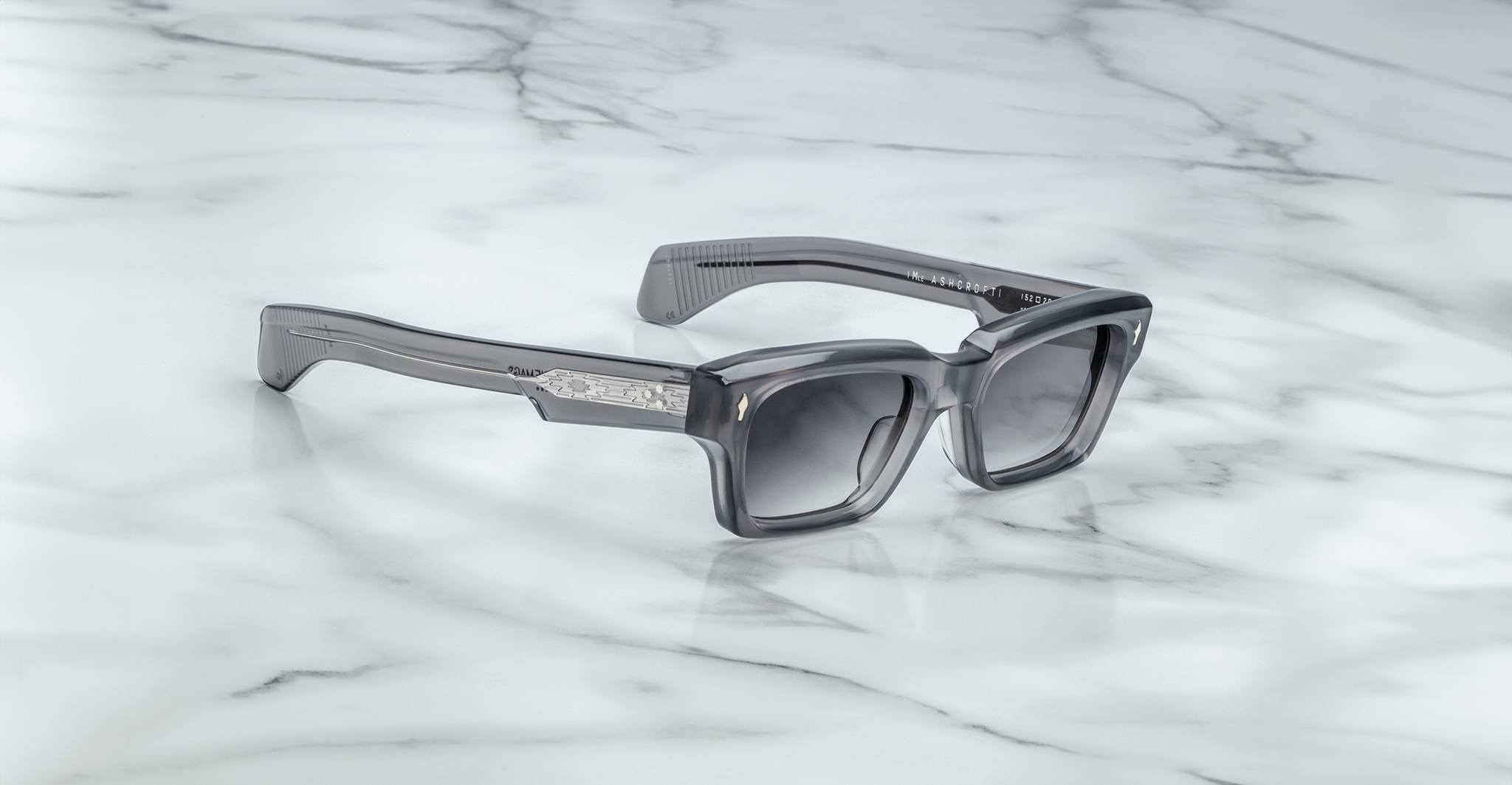 Jacques Marie Mage Ashcroft sunglasses, showcasing gray rectangular thick frames and dark lenses, rest on white marble with gray veining. Metallic accents adorn the arms near the hinges, adding a refined touch.