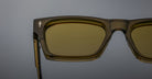 Close-up of the upper left side of Jacques Marie Mage Ashcroft square sunglasses in olive green with yellow-tinted lenses, set against a plain gray background.