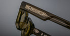 A close-up of the Jacques Marie Mage Ashcroft eyeglasses arm in translucent olive green, showcasing a metallic inlay engraved with lightning bolts and stars, with the glasses’ reflection visible on the surface below.