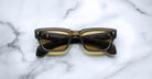 The Jacques Marie Mage Ashcroft sunglasses, featuring rectangular yellow-tinted lenses and dark frames, are displayed face up on a white marble surface.