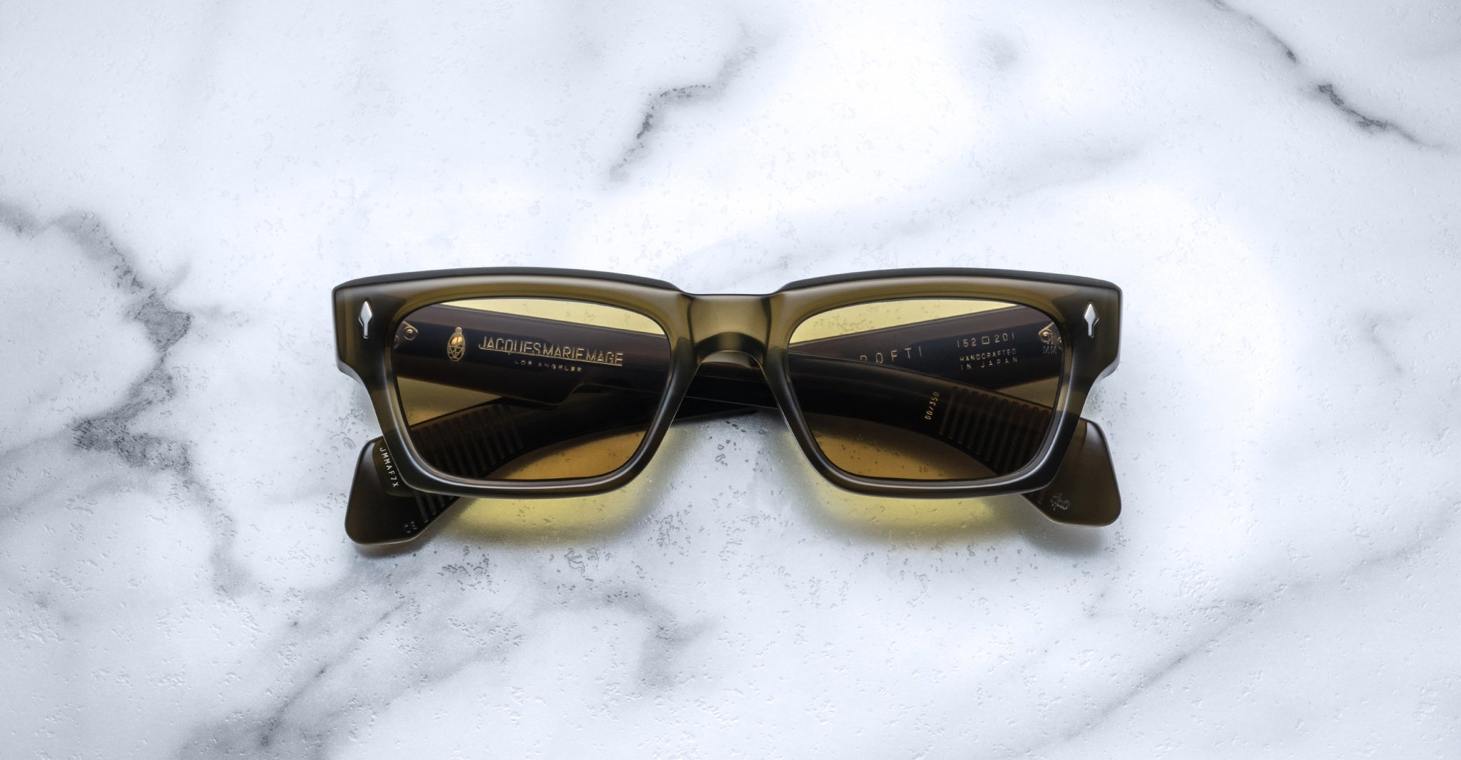 The Jacques Marie Mage Ashcroft sunglasses, featuring rectangular yellow-tinted lenses and dark frames, are displayed face up on a white marble surface.