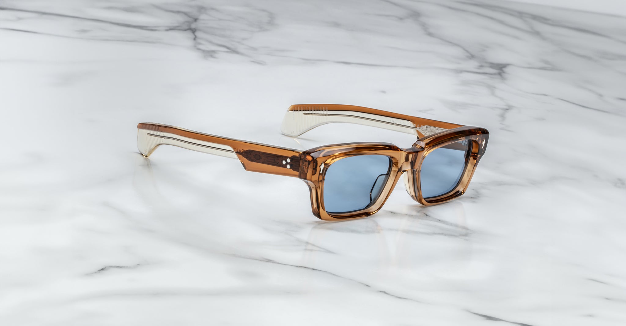 Jacques Marie Mage Ashcroft translucent brown square sunglasses with blue lenses are angled on a white marble surface with grey veining, showcasing both the front and a side arm.