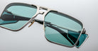 Close-up of Jacques Marie Mage Assen rectangular sunglasses with teal-tinted lenses, a silver metallic frame, and a modern double bridge, resting on a light gray surface.