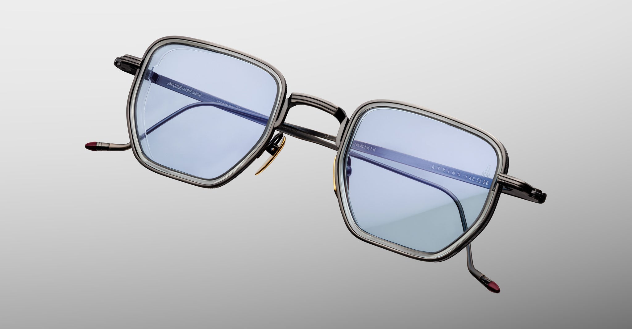 The Jacques Marie Mage Atkins eyewear, featuring large square blue-tinted lenses and a thin dark metal frame, is displayed against a light gray background.
