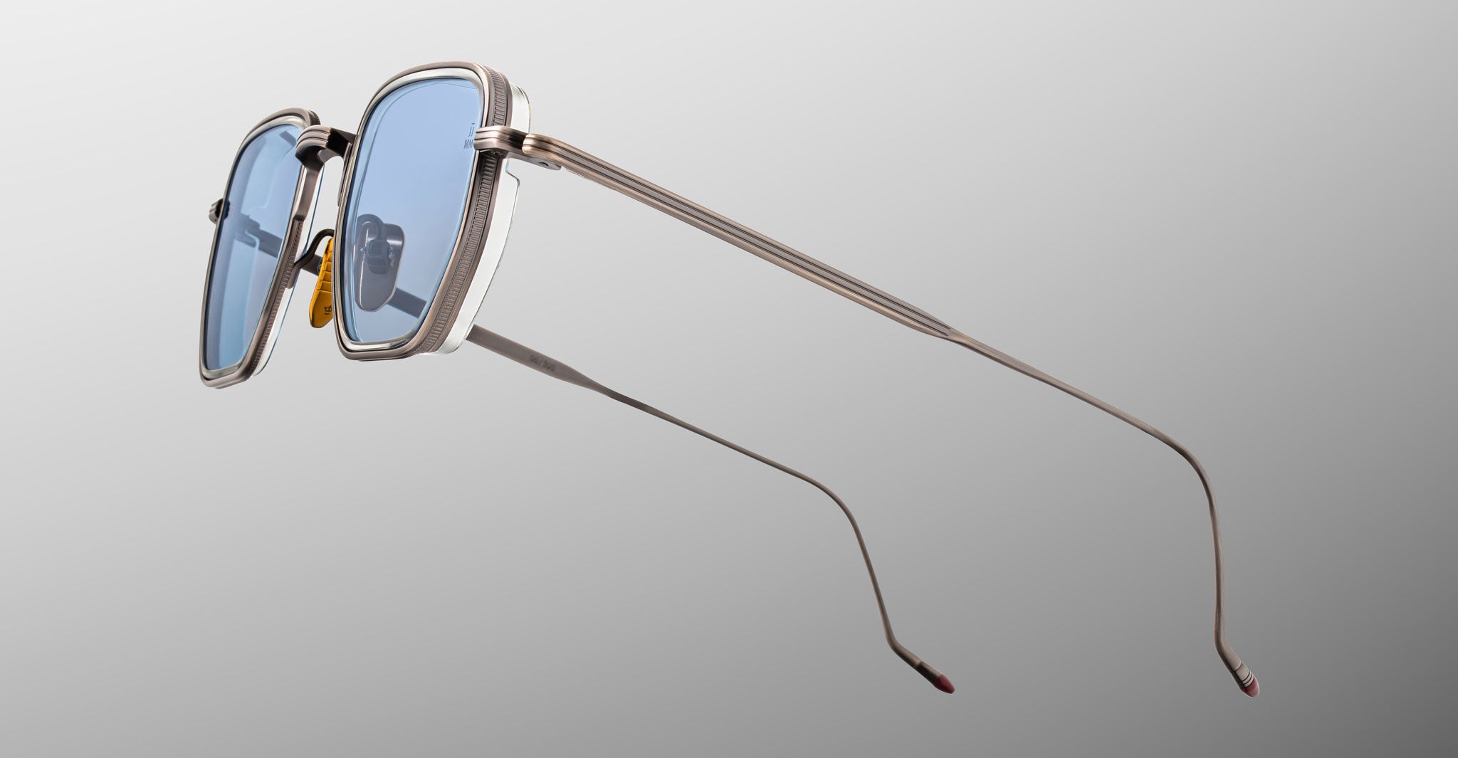 The Jacques Marie Mage Atkins sunglasses feature blue-tinted rectangular lenses, sleek silver metal frames, and slim elongated arms, displayed against a light gray background.