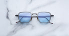 Jacques Marie Mage Atkins eyeglasses from the Titanium Series, with square blue-tinted lenses and slim metal frames, rest on a white marble surface veined in gray.