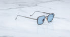 The Jacques Marie Mage Atkins, modern square-shaped sunglasses from the Titanium Series with thin metal frames and blue-tinted lenses, rest elegantly on a white marble surface.