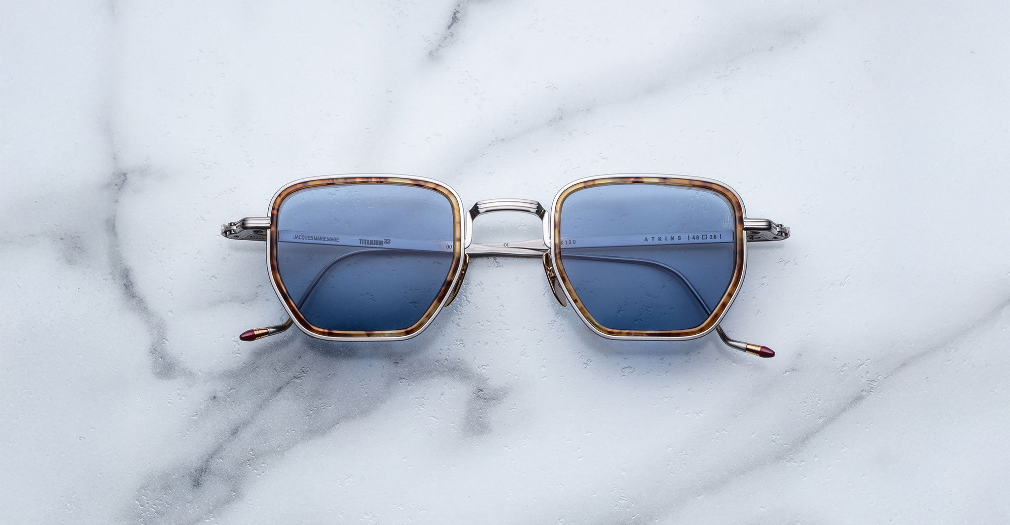 The Jacques Marie Mage Atkins sunglasses, featuring square frames with blue lenses and silver arms, rest on a white marble surface. Water droplets are visible on the lenses.