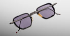 A pair of Jacques Marie Mage Atkins sunglasses featuring a rectangular black frame, purple-tinted lenses, gold nose pads, and thin metal arms is displayed upside down on a light gradient background.