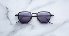 The Jacques Marie Mage Atkins—black, square sunglasses with dark lenses—rest on a white marble surface, showcasing handcrafted Japanese craftsmanship and timeless elegance from a top-down view.