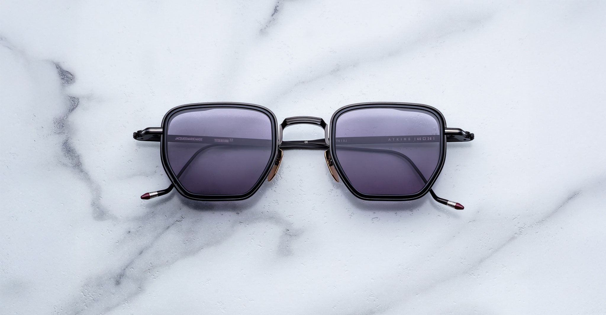 The Jacques Marie Mage Atkins—black, square sunglasses with dark lenses—rest on a white marble surface, showcasing handcrafted Japanese craftsmanship and timeless elegance from a top-down view.