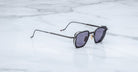 A pair of Jacques Marie Mage Atkins sunglasses with black square frames and dark lenses rests on a white marble surface.
