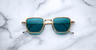 Jacques Marie Mage Atkins square sunglasses with gold frames and blue lenses, viewed from above.